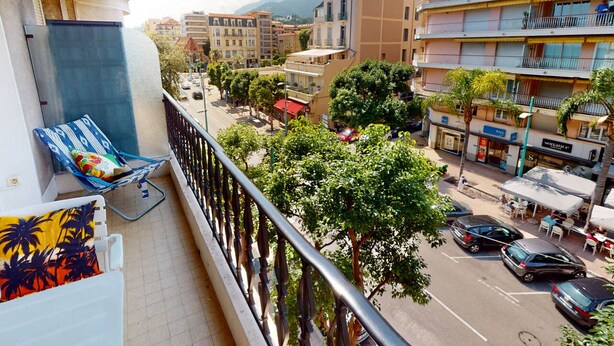 En balkon med en blå stol og en farverig pude, med udsigt over en gade med biler og bygninger.