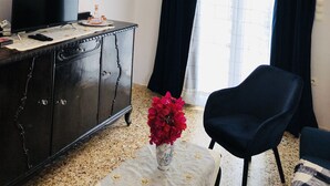 Een kamer met een kroonluchter, een televisie op een kastje, een lijst met een schilderij, een klein fotolijstje, een vaas met rode bloemen en een donkerblauwe stoel.