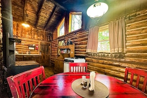 A rustic cabin interior with a wooden dining table, red chairs, a kitchen area with a stove, and a large window.