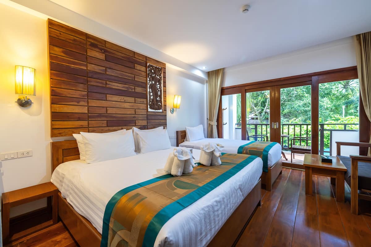 A hotel room with two beds, wooden headboard, large windows, and a balcony with a view of greenery.