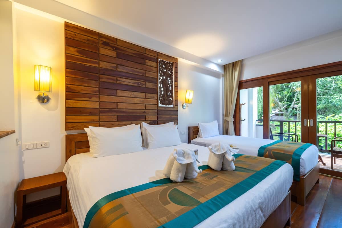 A hotel room with two beds, wooden headboard, wall-mounted lights, and a view of greenery through large windows.