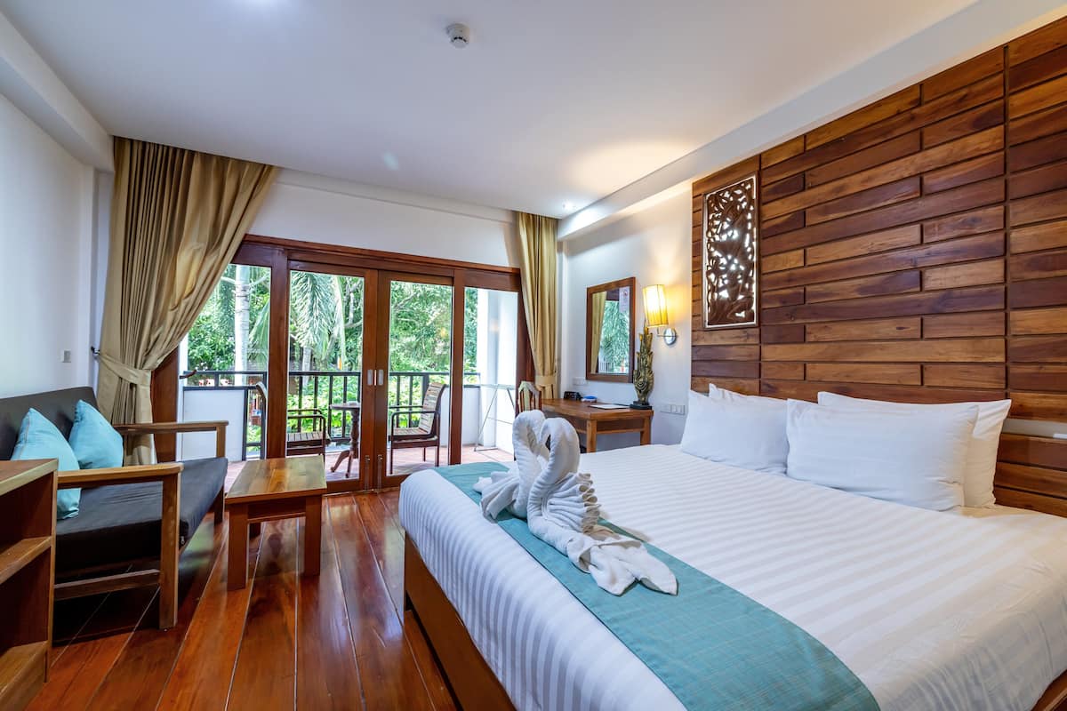 A hotel room with a wooden headboard, a large bed with white linens, a wooden desk, a chair, and a balcony with a view of greenery.