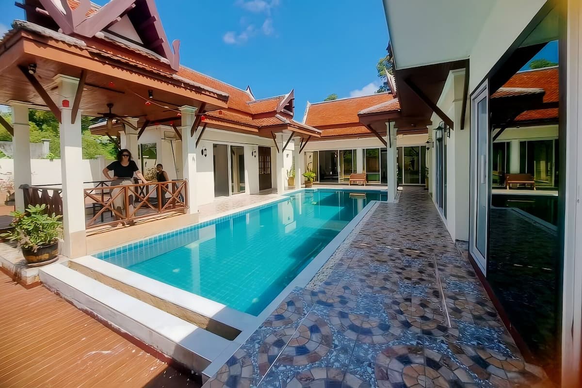 A swimming pool with a tiled deck, a covered seating area, and a building with a tiled roof.