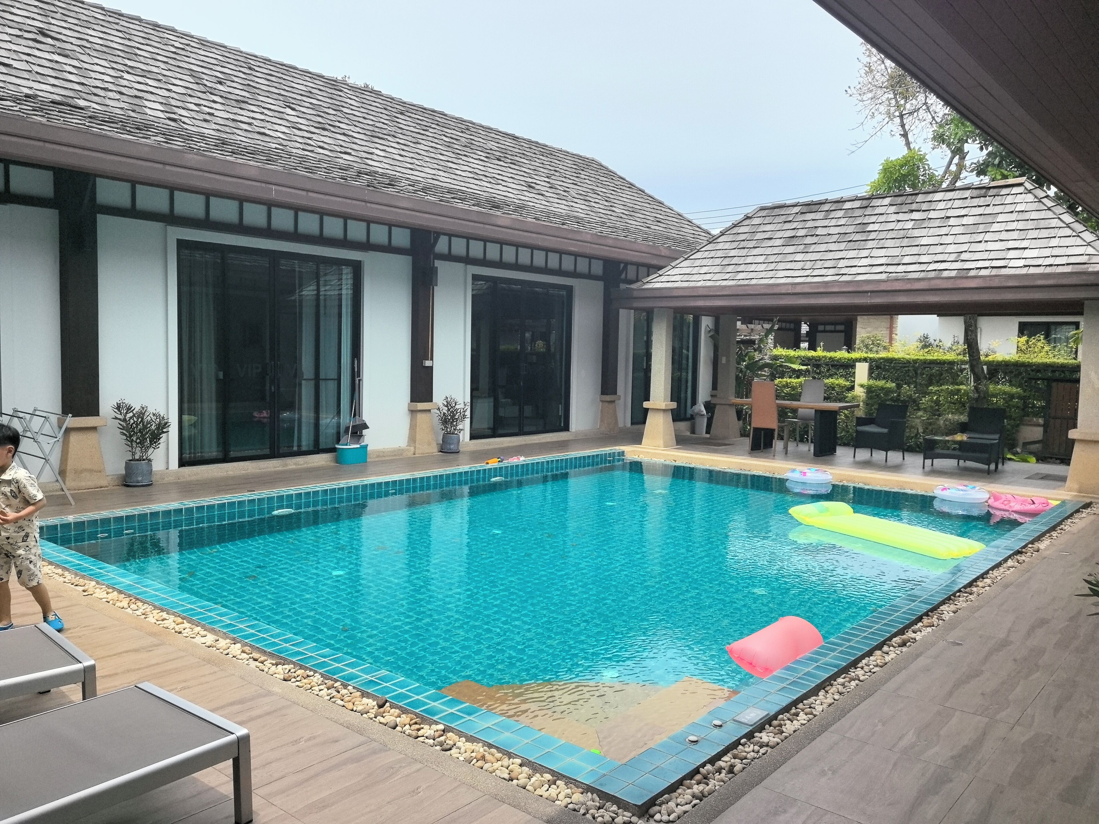 Una piscina con bordes de azulejos, un área de descanso con mesa y sillas, y un porche cubierto con mobiliario exterior.