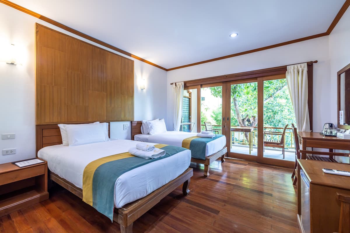 A hotel room with two beds, wooden flooring, a balcony with a view, and a desk with a chair.