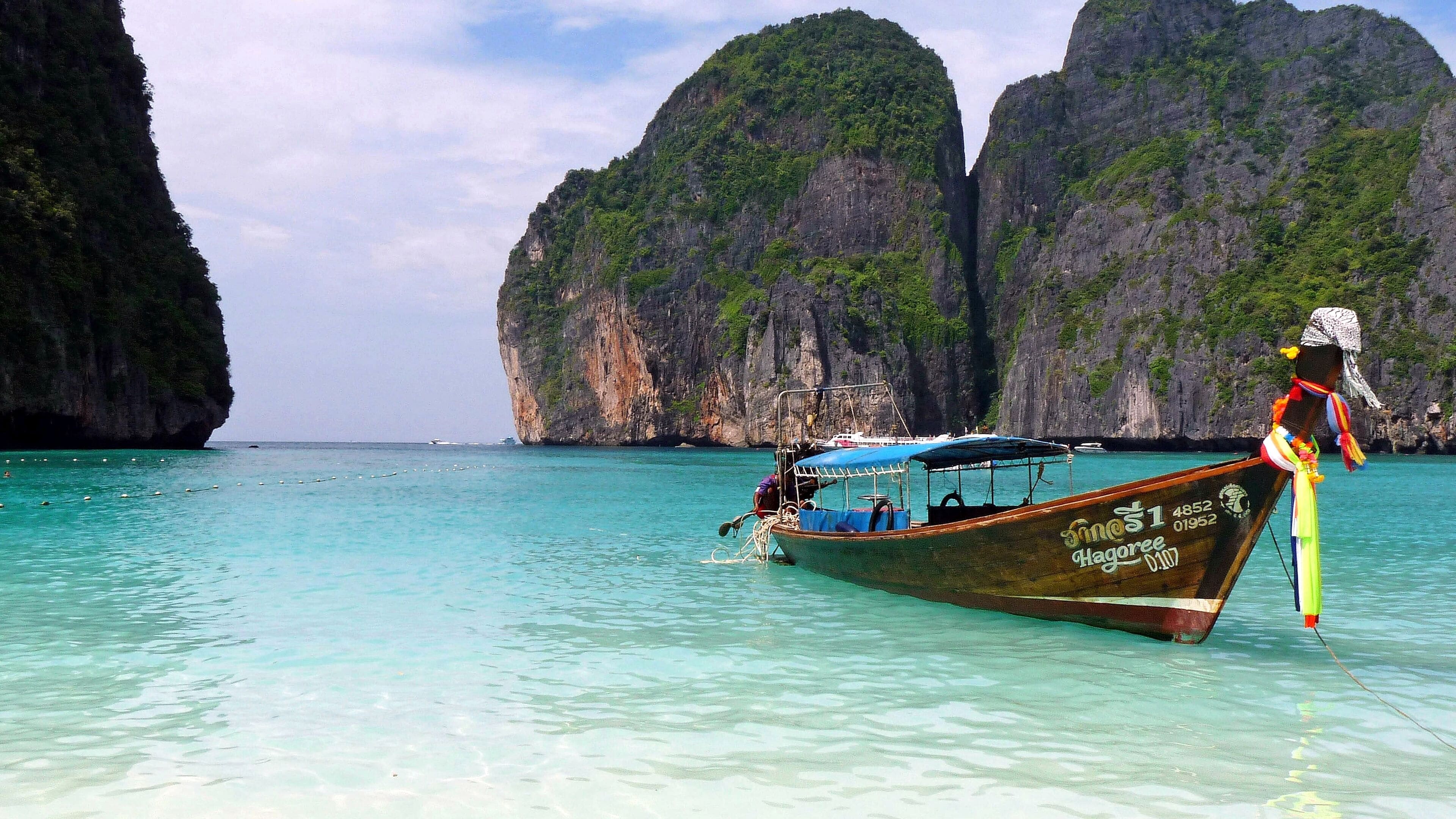 A boat with a person standing on the bow, a clear turquoise sea, and large limestone cliffs in the background.