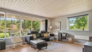 Ein modernes Wohnzimmer mit einer schwarzen Ledersofa, einem weißen Schaukelstuhl und einem Essbereich mit Tisch und Stühlen. Große Fenster bieten Ausblick ins Freie.