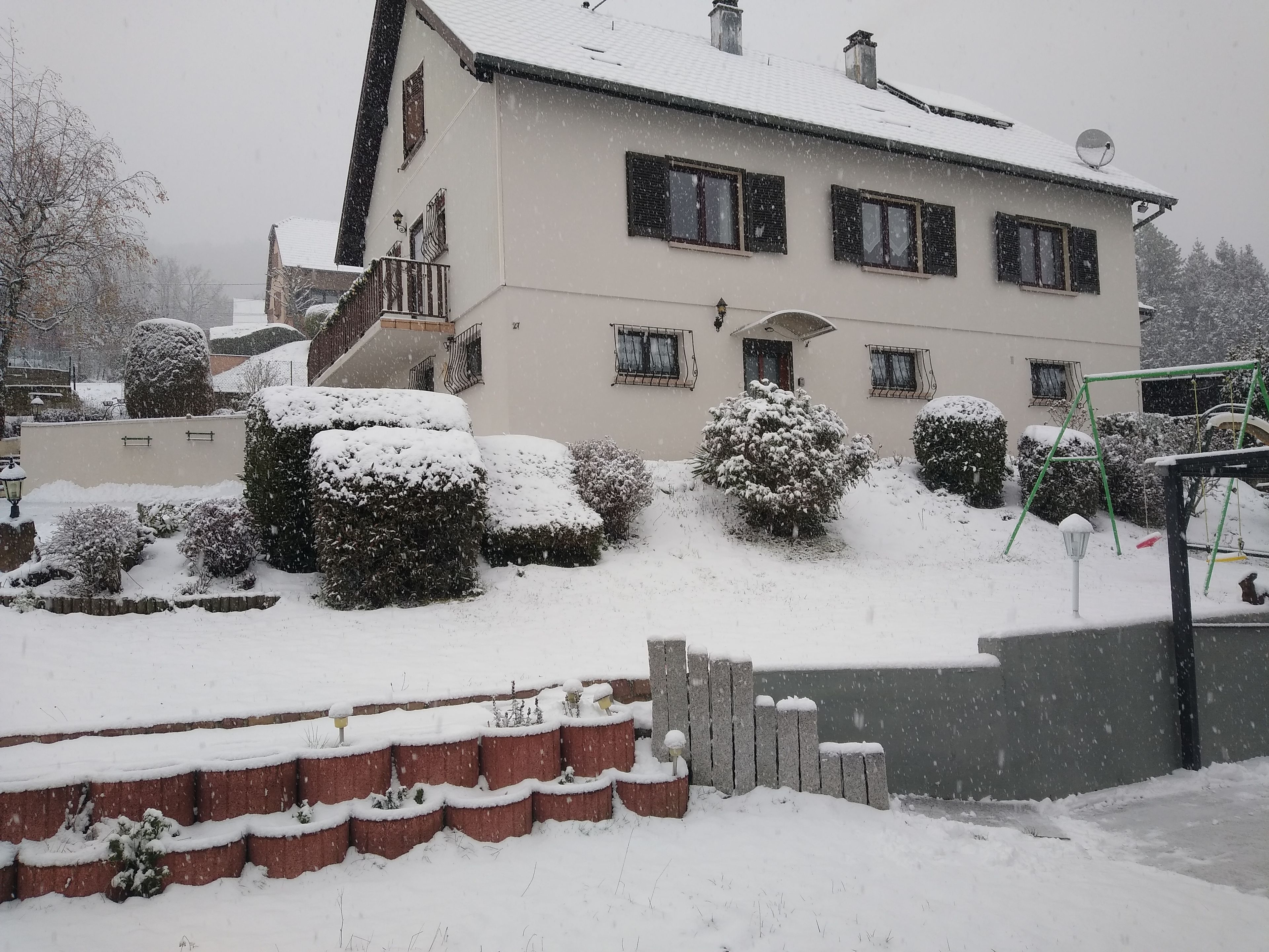 Edifício residencial coberto de neve com antena parabólica, balanço e arbustos com neve.