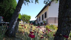 Ein Garten mit roten Blumen und Bäumen, ein weißes Gebäude mit Fenstern und ein klarer Himmel.