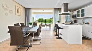 A modern kitchen with a central island, a dining area with a long table and chairs, and a view of greenery outside. - Moonlight Villa | Harbour Lakes at Palmas del Mar (Palmas Del Mar)