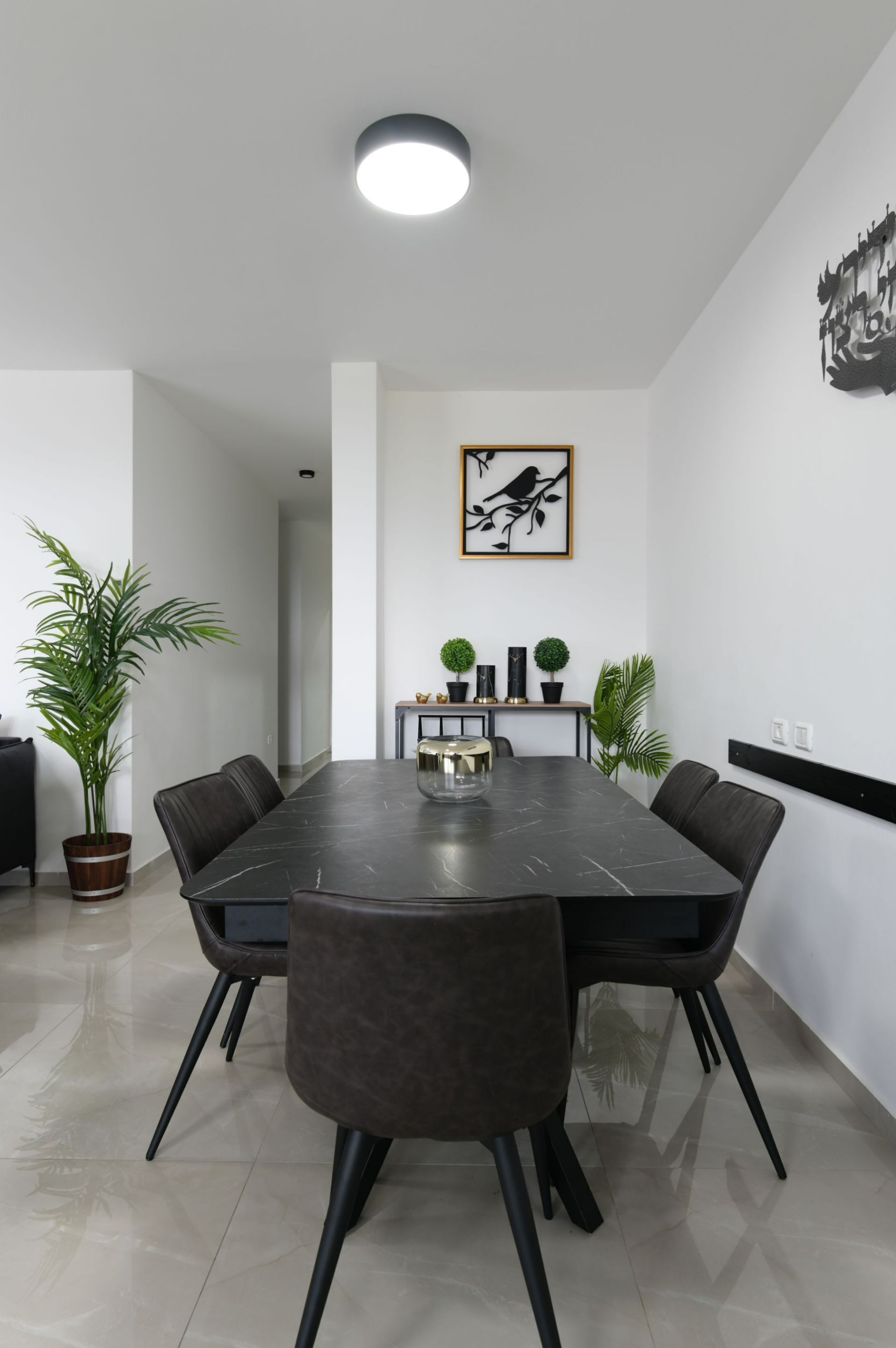 Ein moderner Essbereich mit einem rechteckigen Tisch, sechs Stühlen und einer Deckenleuchte. Es gibt Kübelpflanzen und ein gerahmtes Kunstwerk an der Wand.