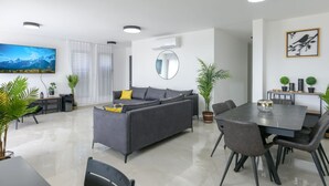A modern living room with a gray sofa, dining table, and chairs. There is a large flat-screen TV mounted on the wall, a round mirror, and several potted plants. - YalaRent Villa lumièra with Private Pool (Migdal)