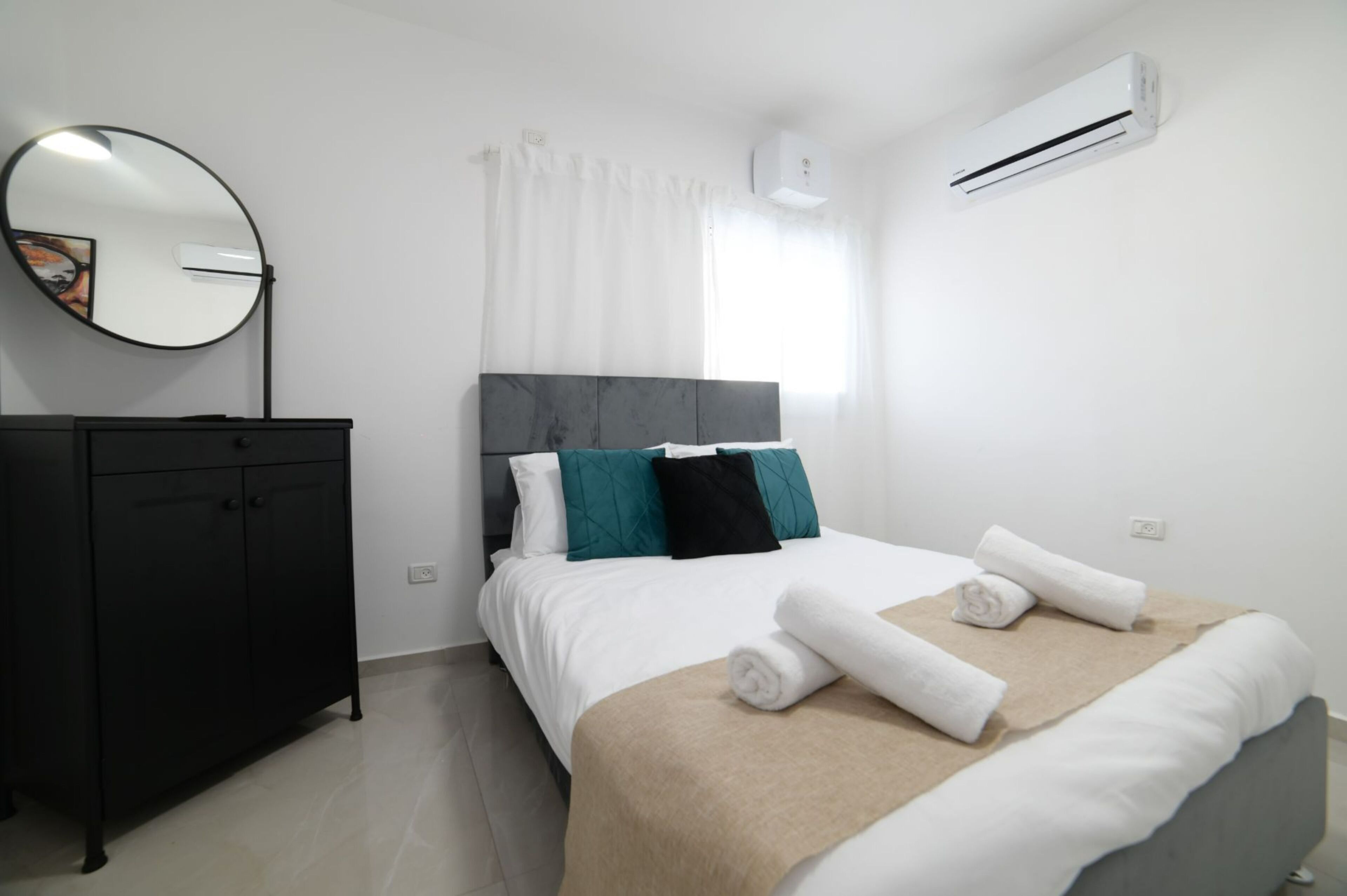 A neatly arranged bedroom with a bed, a dresser, a round mirror, and an air conditioning unit.