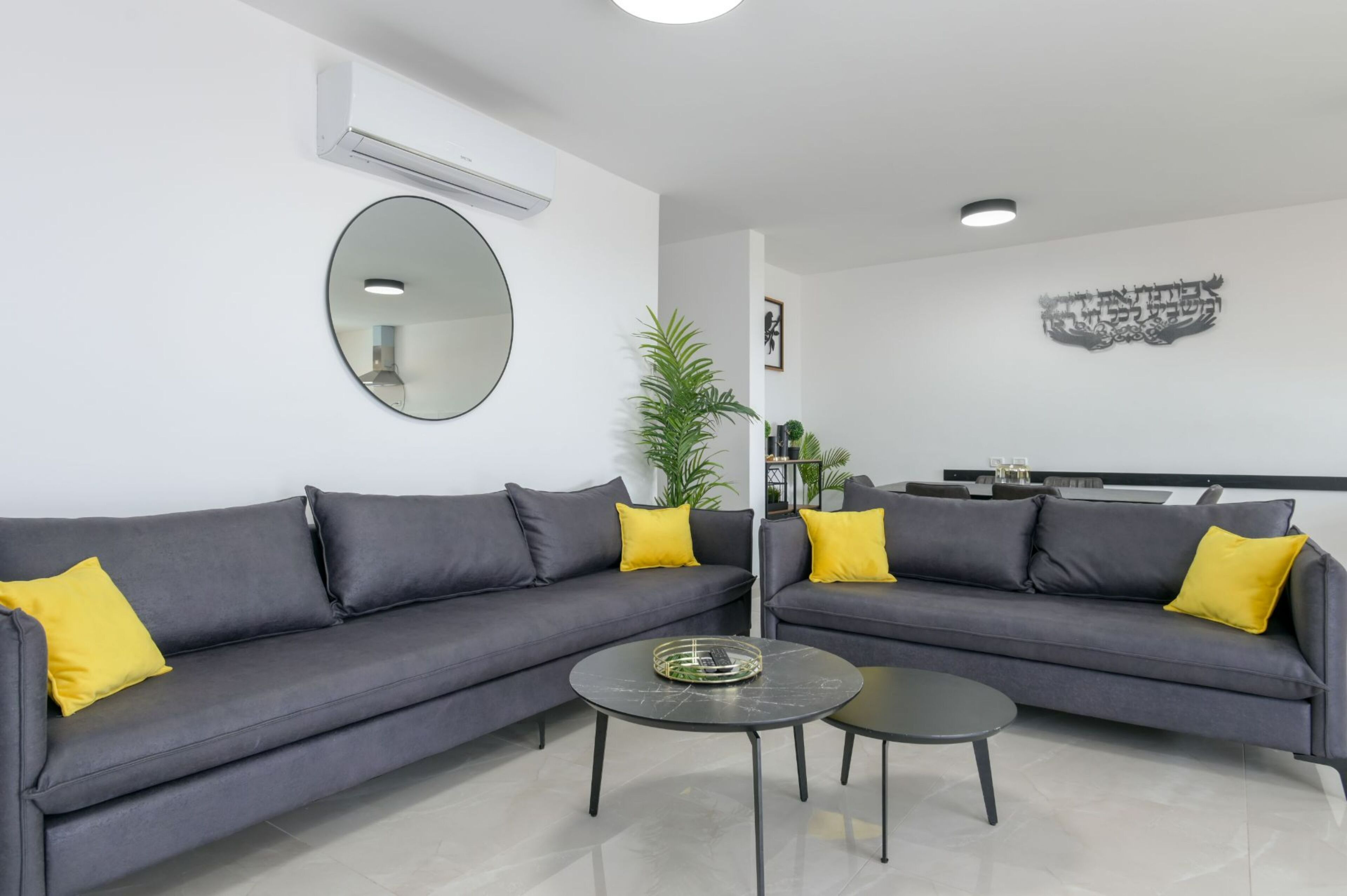 A modern living room with two grey sofas, each with a yellow pillow, and a round mirror on the wall. There is a small round table with a tray in the center.