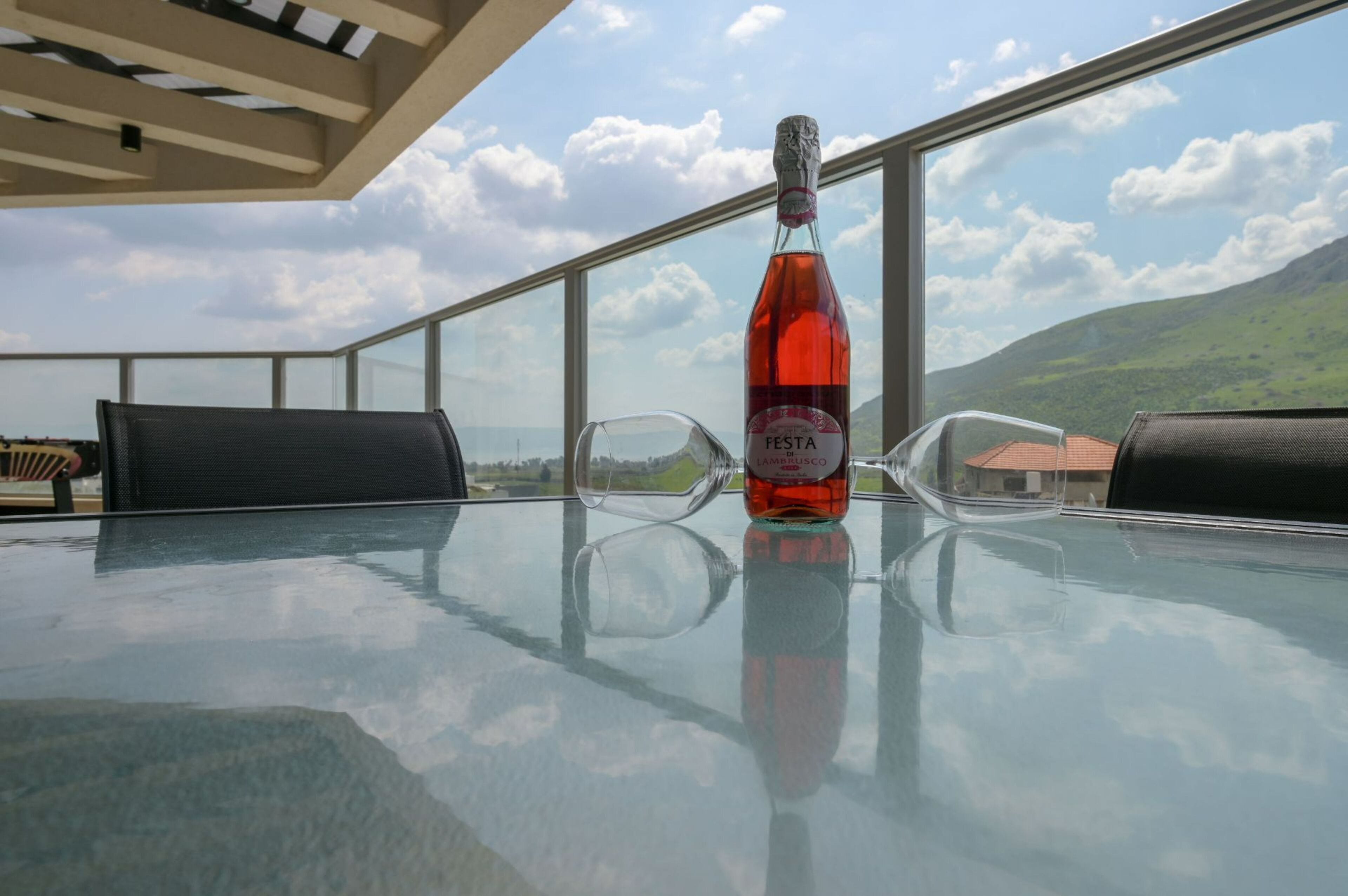 A glass table with a bottle of Festa Lambisco wine, two glasses, and black chairs on a balcony with a view of mountains and sky.