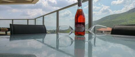 A glass table with a bottle of Festa Lambisco wine, two glasses, and black chairs on a balcony with a view of mountains and sky.