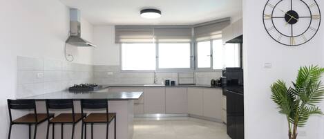 A modern kitchen with a central island, bar stools, a large window, and a wall-mounted clock.