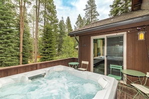 A hot tub on a wooden deck with a glass door leading to a covered area, surrounded by trees.