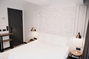Habitación de hotel con cama, mesita de noche, lámpara y termostato en la pared.