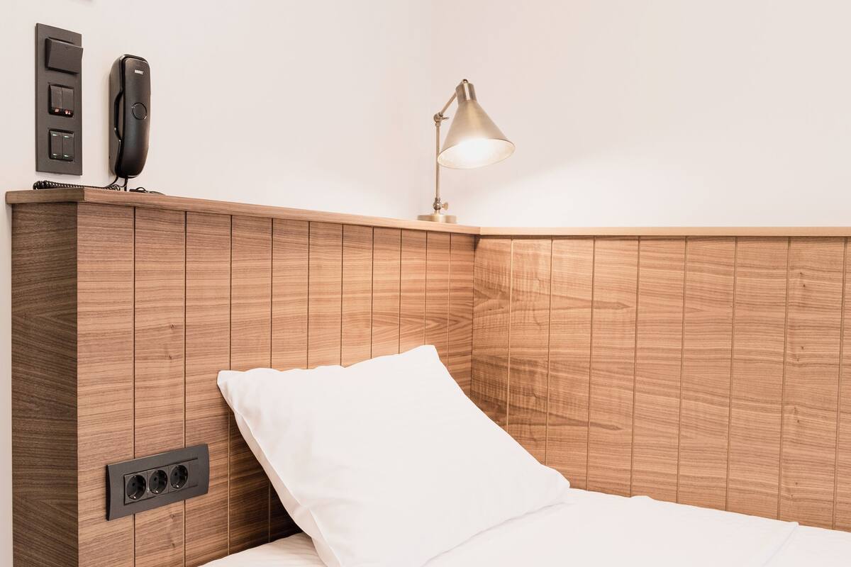 A neatly made bed with a wooden headboard, a bedside lamp, and a thermostat on the wall.