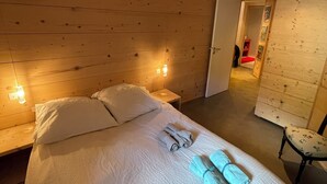 A bedroom with a wooden wall, a bed with white bedding, two bedside lamps, and a chair with a floral cushion. - Chalet au Sud, Face aux Glaciers (Tignes)