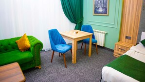 Una stanza con un divano verde, due poltrone blu, un tavolo in legno, un letto con copriletto verde, un comodino e un quadro appeso al muro.
