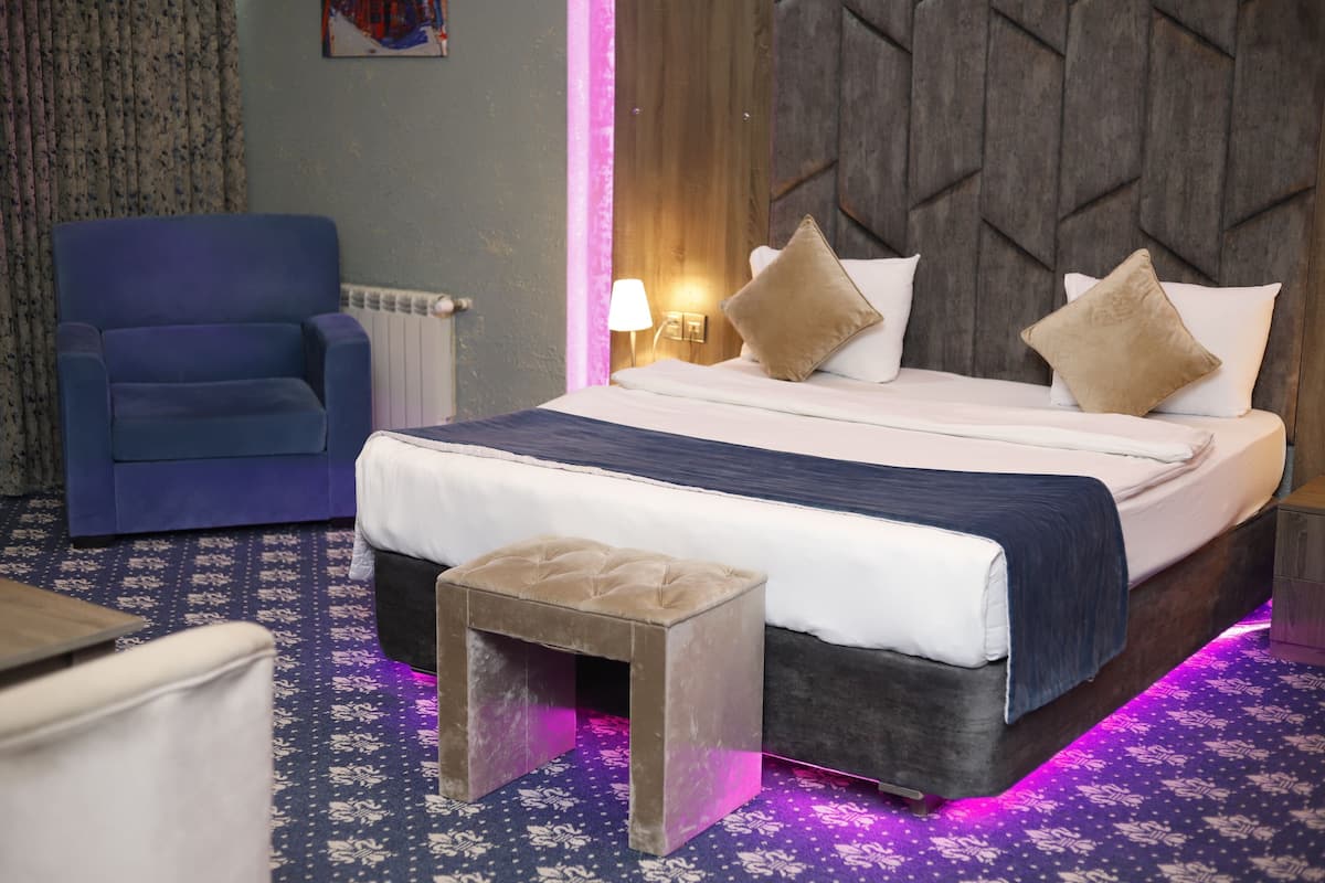 Une chambre d’hôtel avec un lit, une table de chevet, une lampe, une chaise et un petit tabouret.