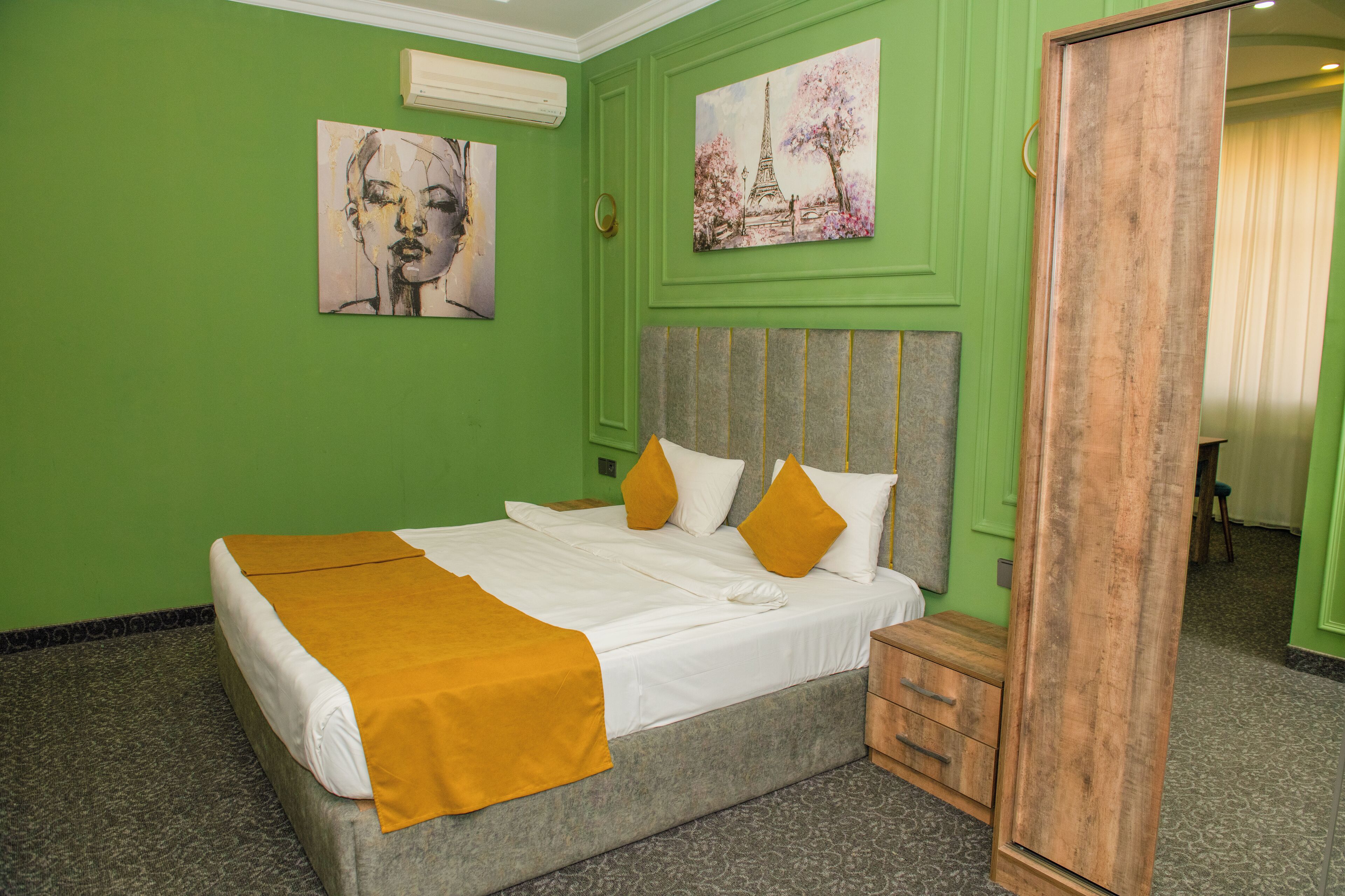 A neatly made bed with a white and yellow bedspread, a grey headboard, and two yellow pillows. There is a wooden nightstand with drawers, a green wall with two paintings, and a wooden wardrobe.