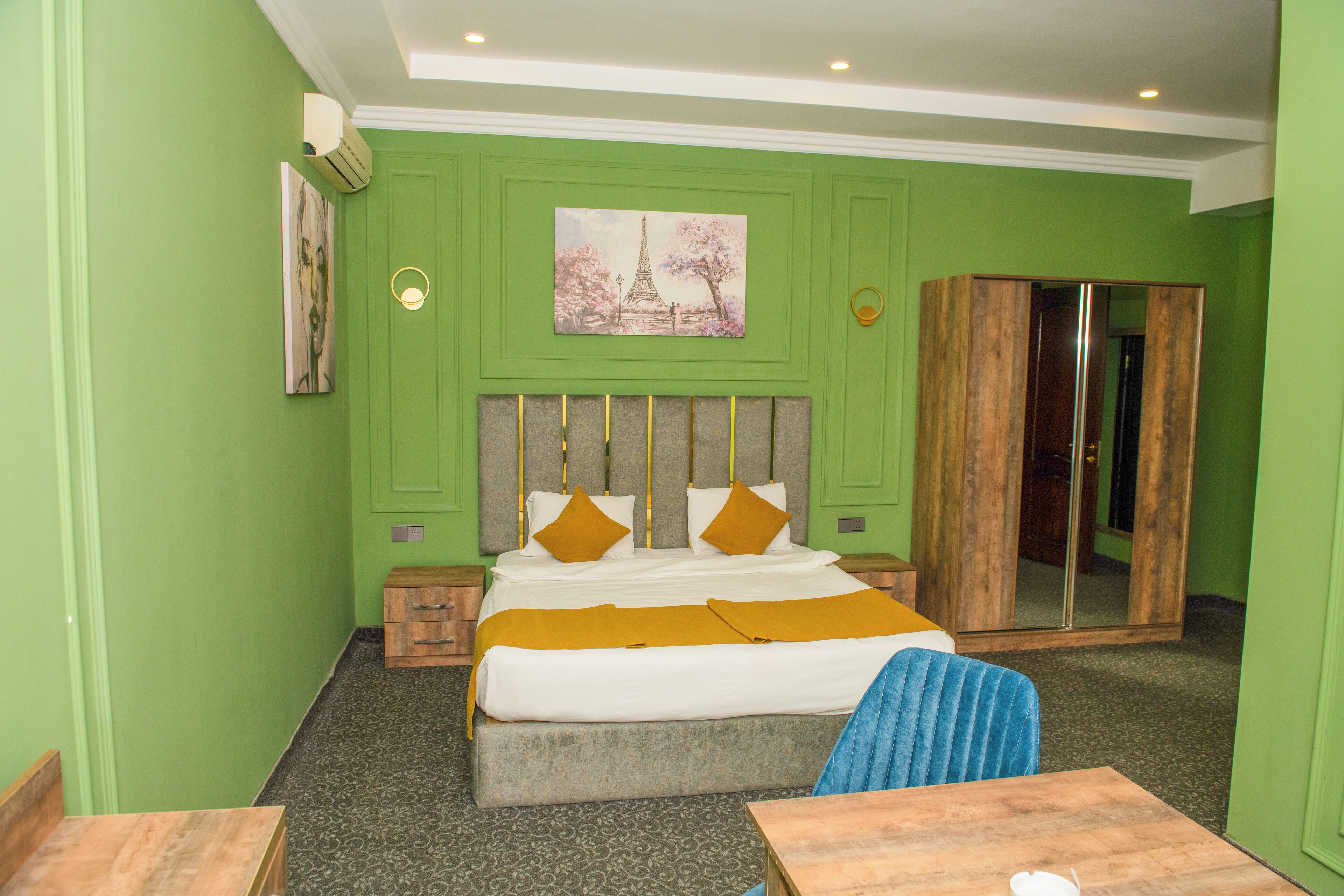 A hotel room with a bed, a desk, a chair, a wardrobe, and a painting on the wall.