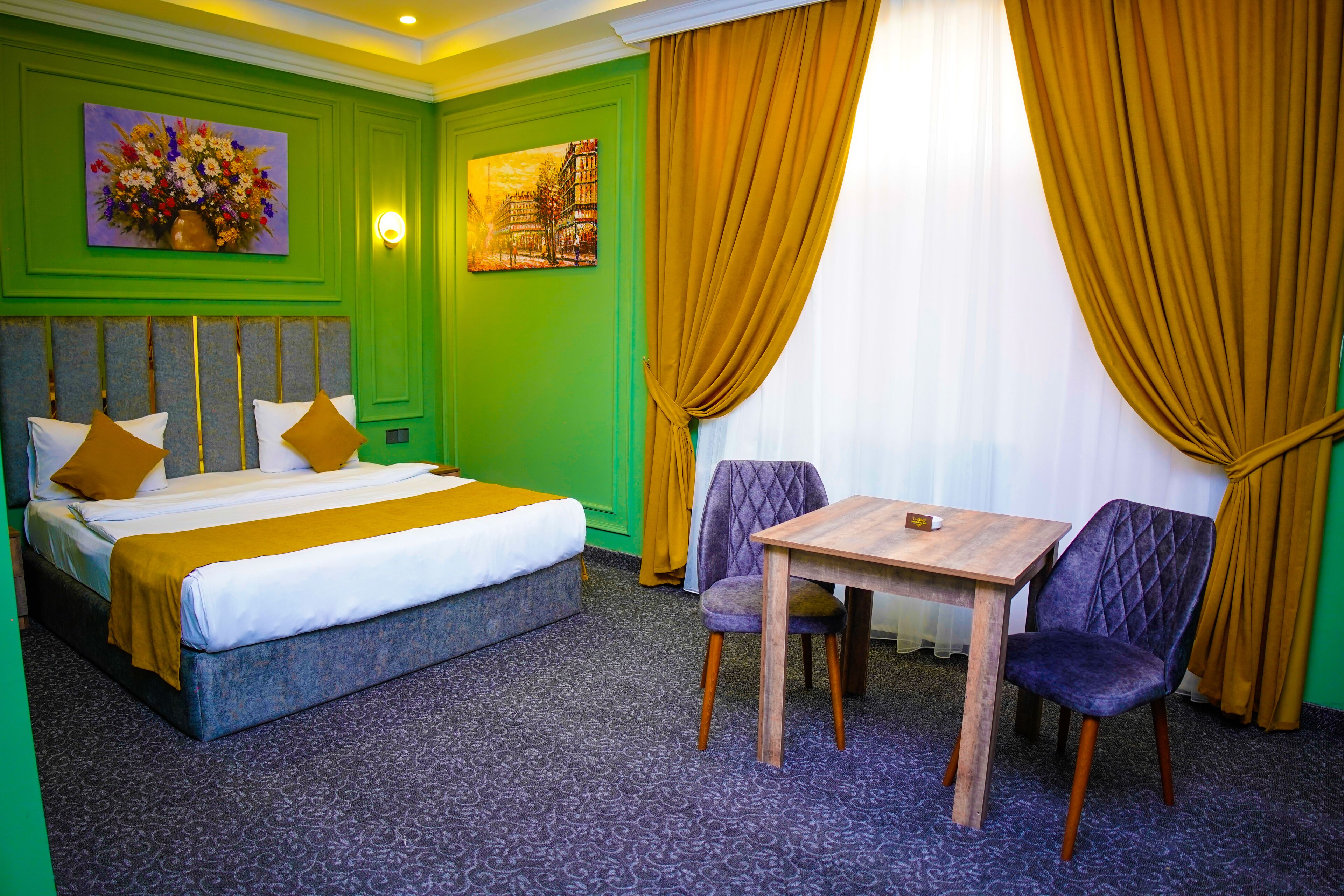 A hotel room with a bed, a table, two chairs, a painting, and a window with curtains.
