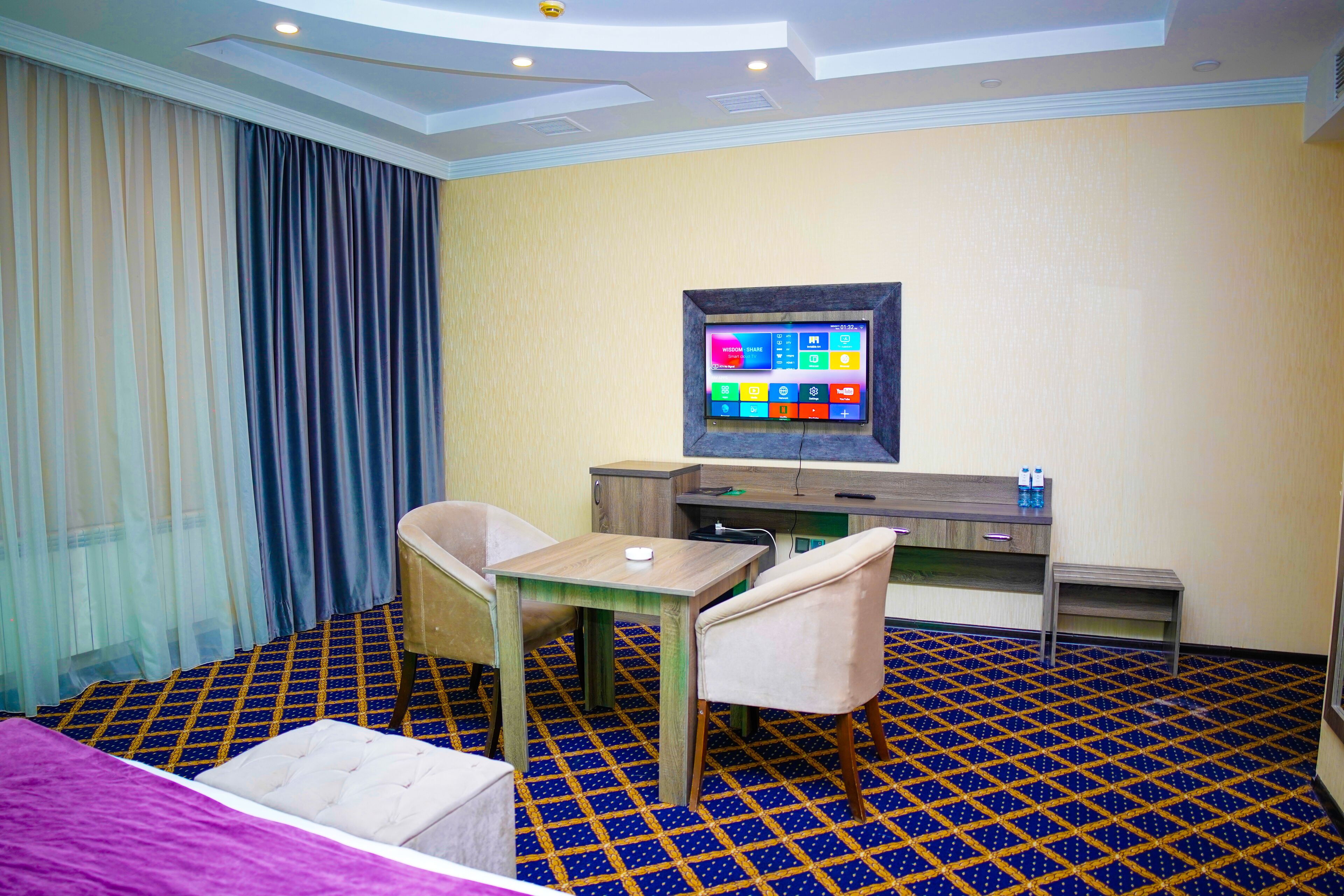 A hotel room with a bed, a desk with a computer, two chairs, a small table, and a large window with curtains.