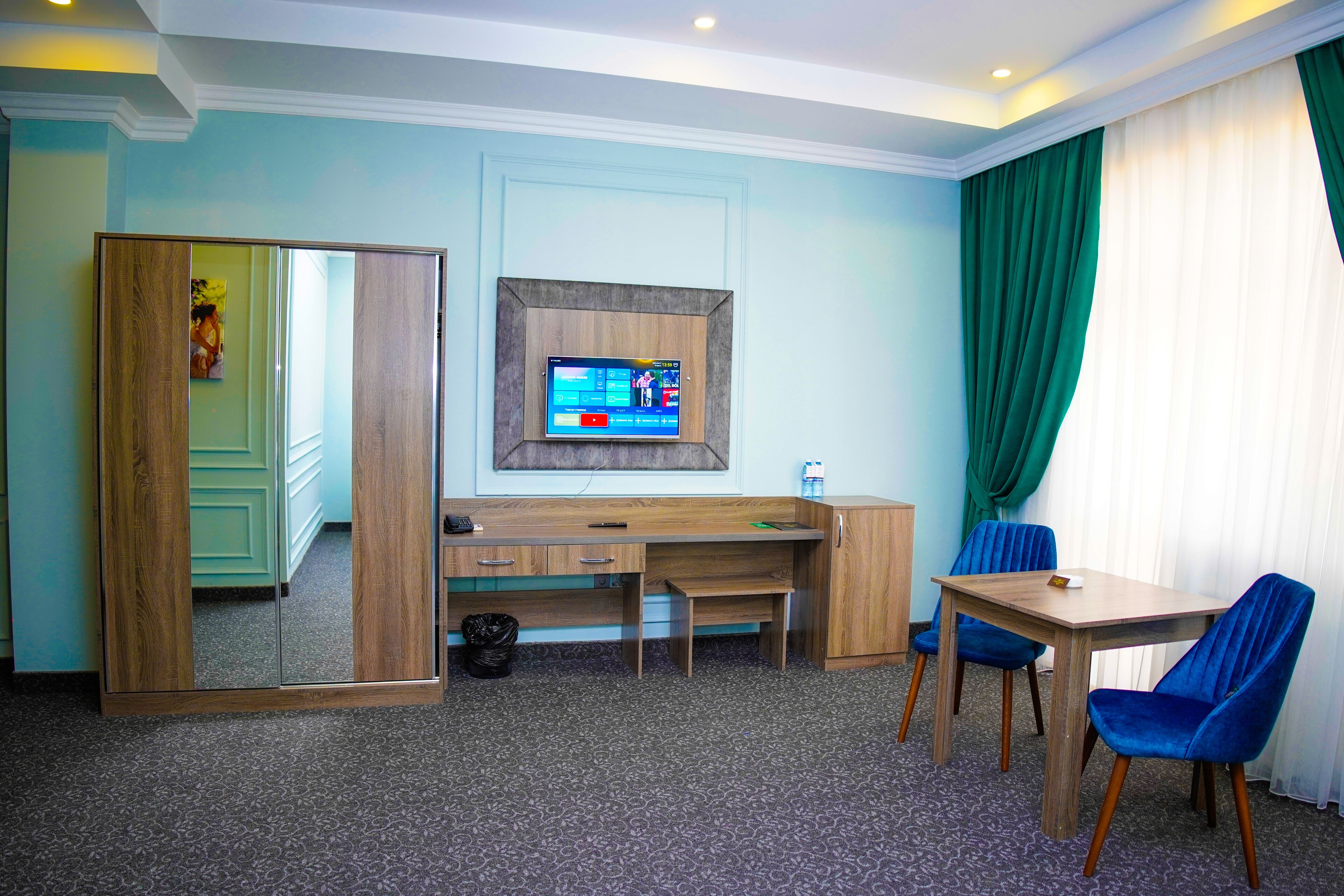 A hotel room with a wooden desk, two blue chairs, a TV on the wall, and a large mirror.