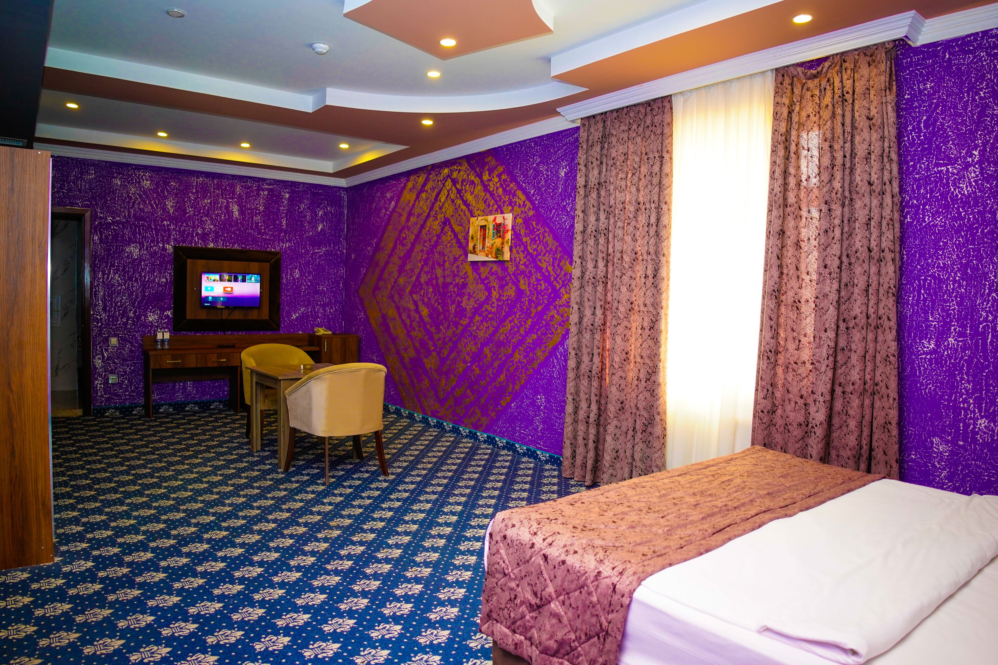 Ein Hotelzimmer mit violettem Wandmuster, einem Bett mit rosa Bettdecke, einem Schreibtisch mit Stuhl und einem an der Wand befestigten Fernseher.