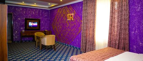 Ein Hotelzimmer mit violettem Wandmuster, einem Bett mit rosa Bettdecke, einem Schreibtisch mit Stuhl und einem an der Wand befestigten Fernseher.