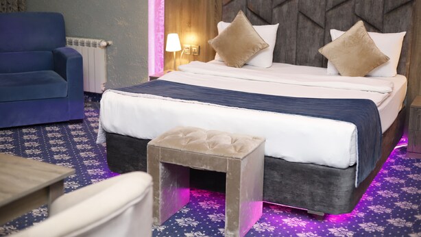 Una camera d'albergo con un letto, una poltrona blu, uno sgabello e una lampada.