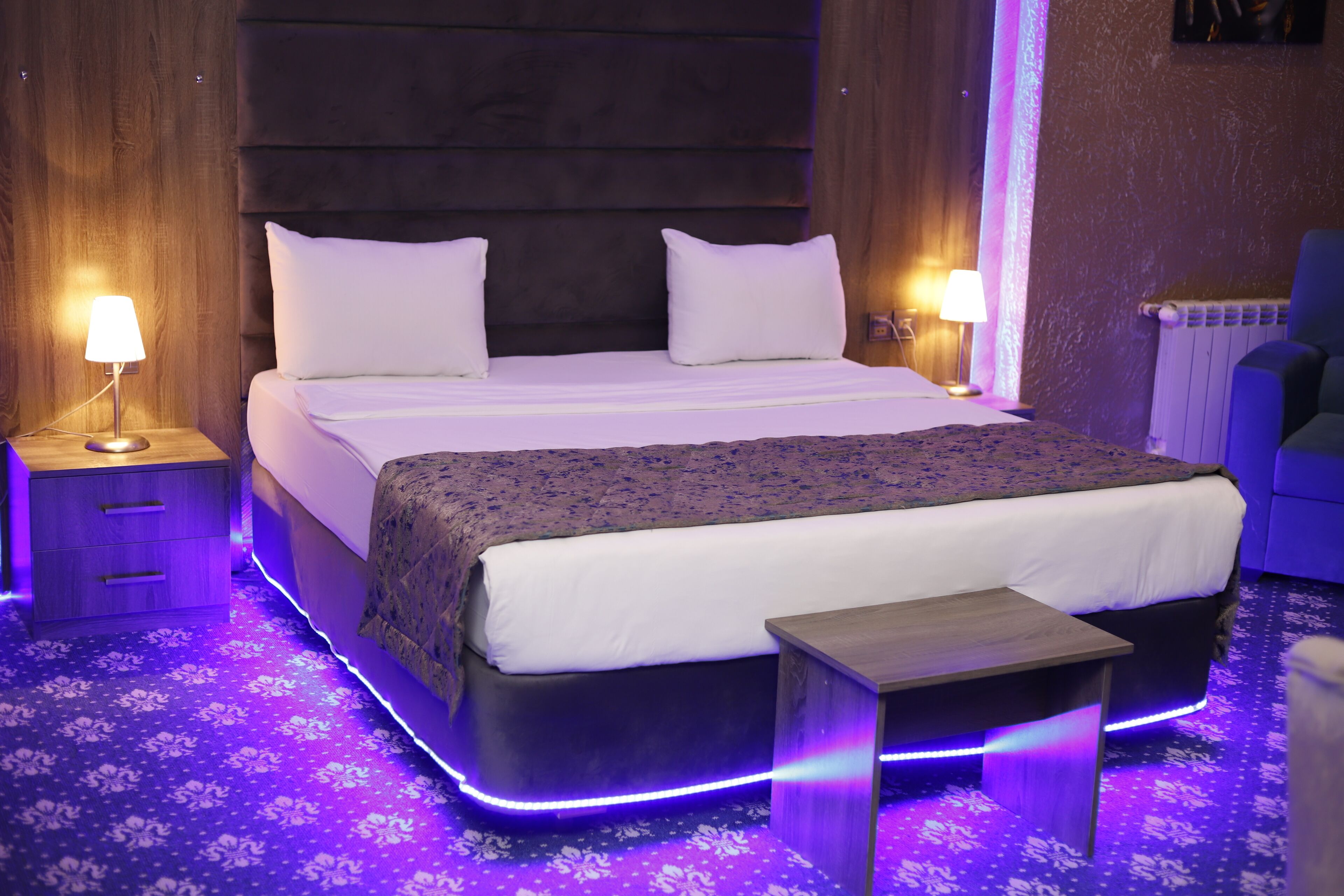 A modern hotel room with a large bed, bedside lamps, a small table, and a blue chair.