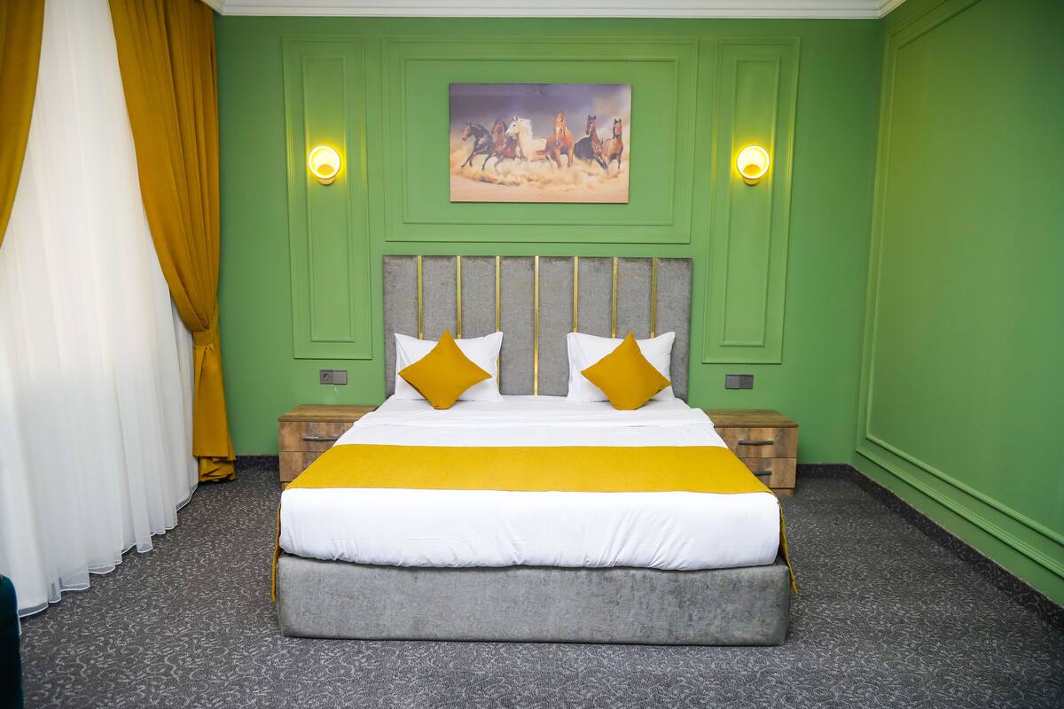 Un lit bien fait, avec une literie blanche et jaune, un mur vert orné d’un tableau, et une table de chevet en bois avec une lampe.