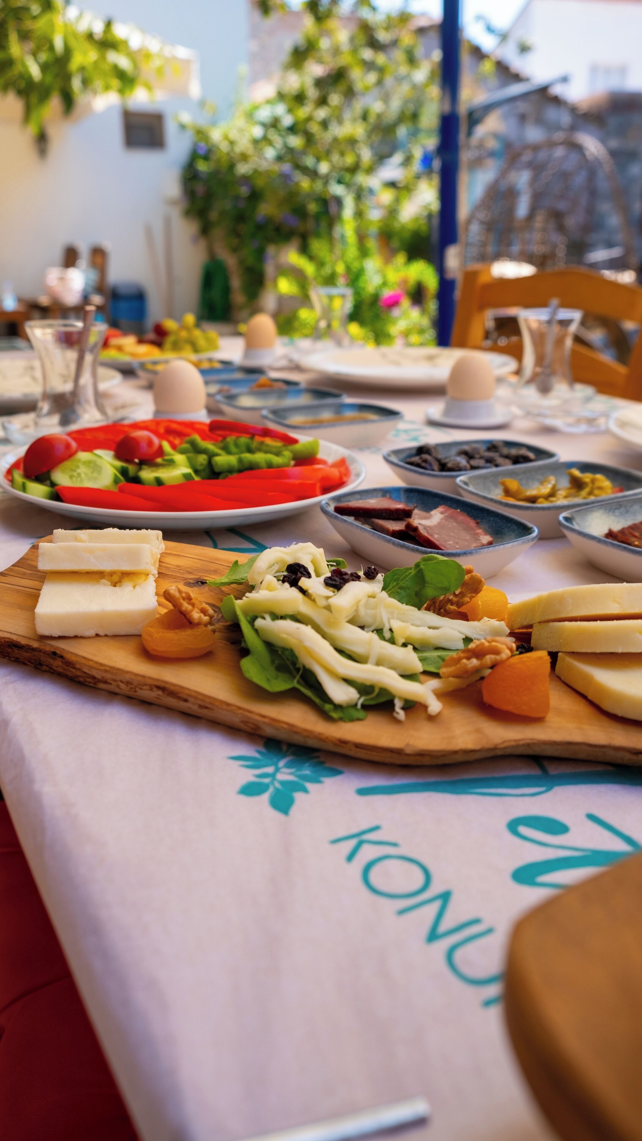 Ein Tisch ist reichhaltig mit Speisen gedeckt, darunter Käse, Nüsse und Gemüse, in einem hellen, offenen Essbereich mit Grünpflanzen im Hintergrund.