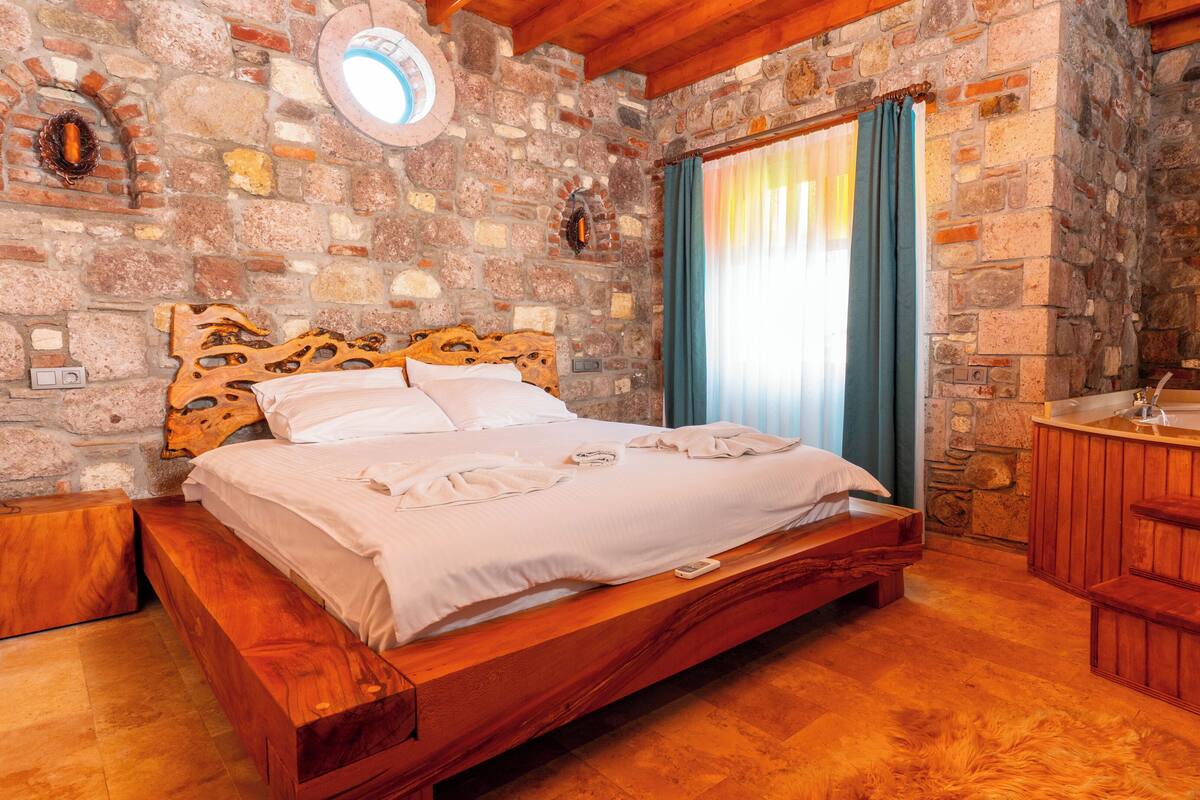Una cama de madera con cabecera, una pared de piedra, un banco de madera y un lavamanos de madera.