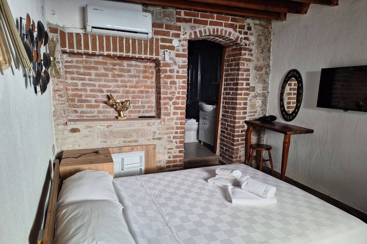 Un dormitorio con edredón a cuadros, una mesita de noche de madera, aire acondicionado instalado en la pared, pared de ladrillo, un espejo y un televisor.