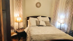 Una cama bien hecha con una colcha blanca y varias almohadas, una mesita de noche con una lámpara y una silla.