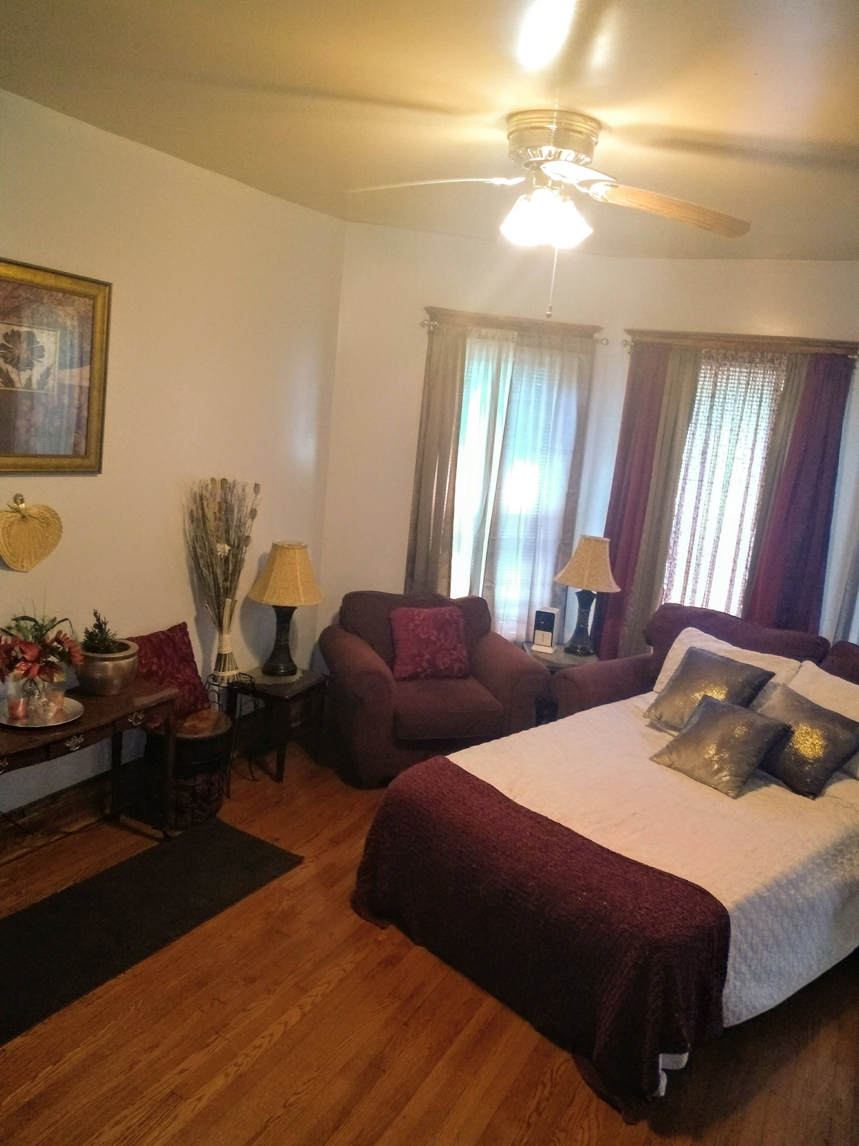 A well-lit bedroom with a large bed, two armchairs, a small table, and a ceiling fan.