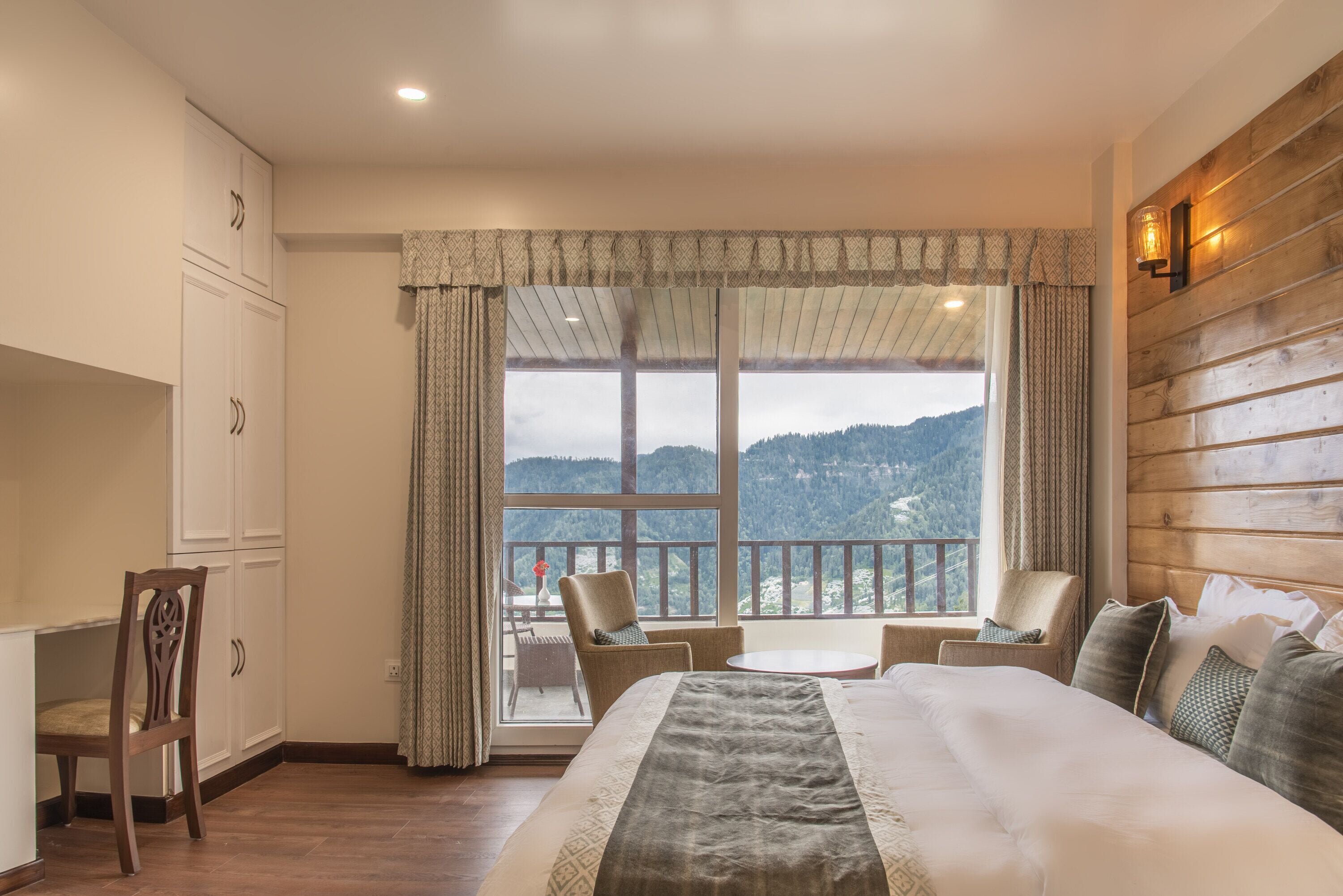 A bedroom with a large bed, a desk, and a chair. There is a balcony with a view of the mountains.