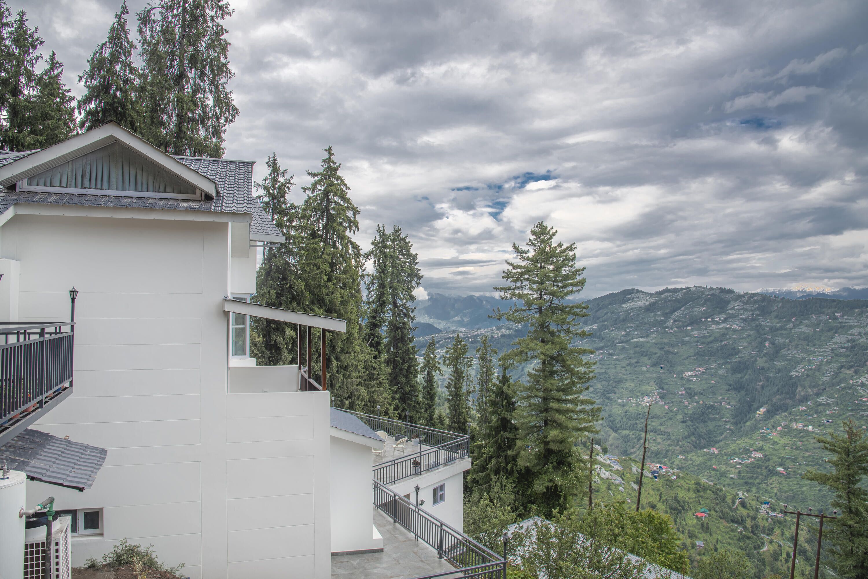 Un edificio moderno blanco con balcón, rodeado de árboles altos y un paisaje montañoso.