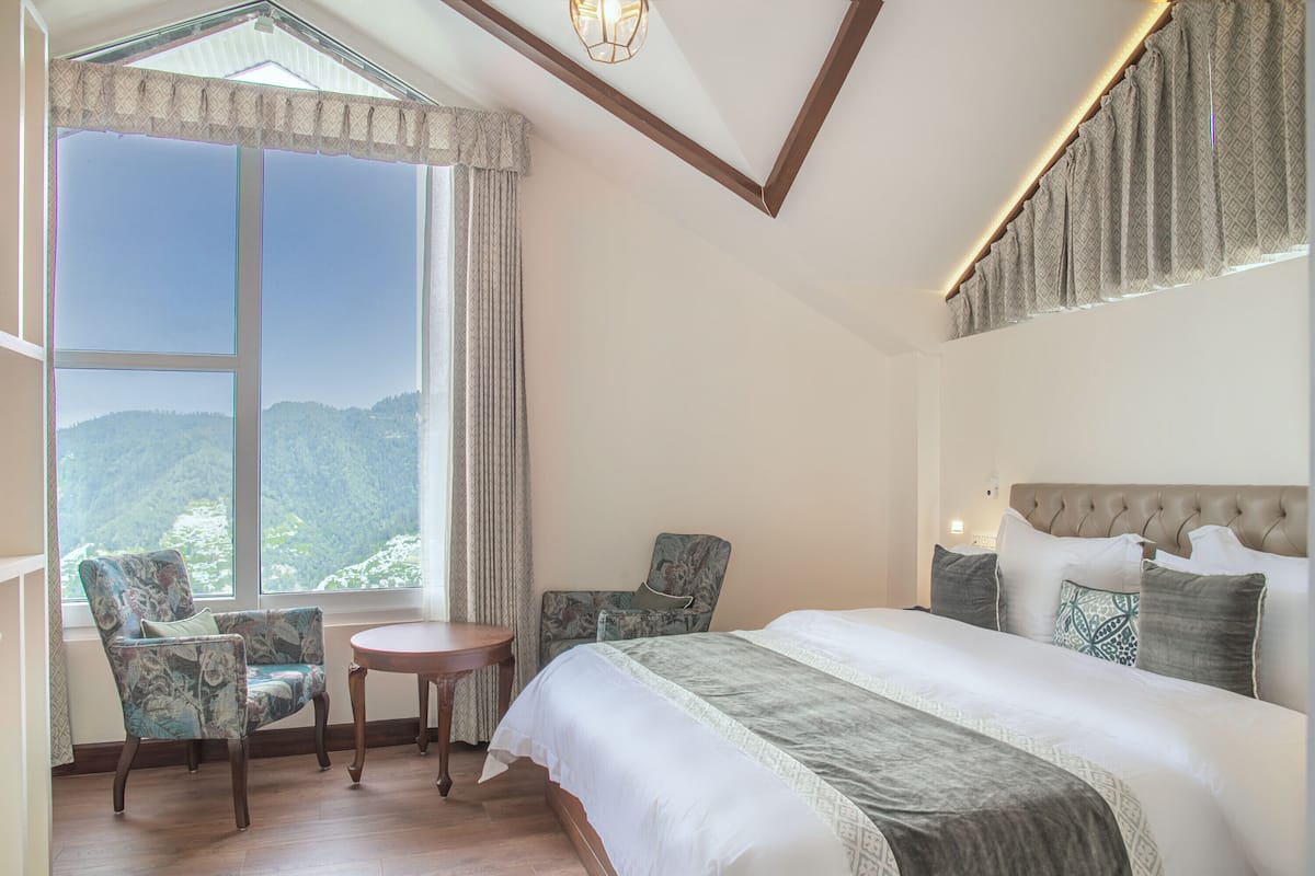 Ein Schlafzimmer mit einem großen Bett, zwei Sesseln, einem kleinen Tisch und Blick auf eine gebirgige Landschaft durch ein großes Fenster.