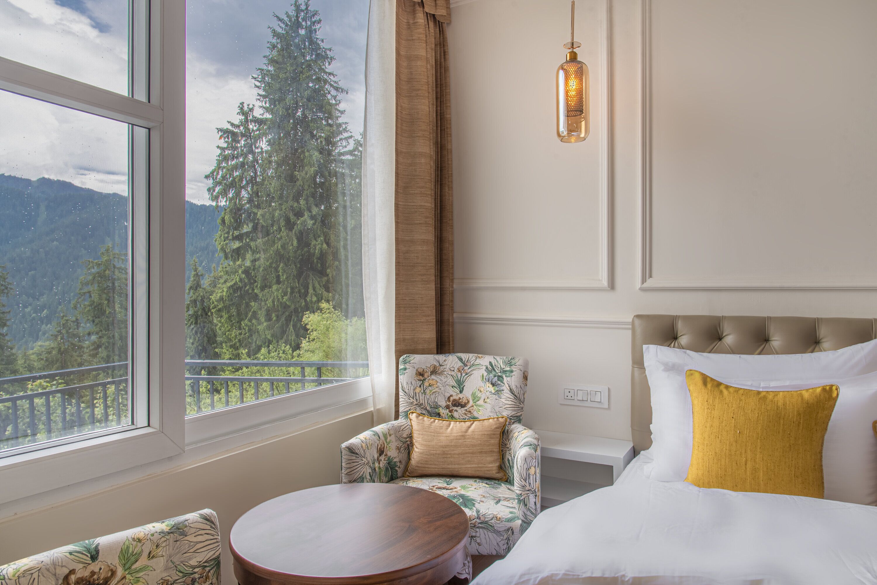 Una habitación de hotel con un ventanal grande, un sillón tapizado con flores, una mesa redonda de madera y una cama con ropa de cama blanca y dorada.