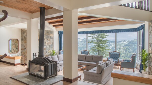 A spacious living room with a stone fireplace, comfortable seating, and a large window offering a scenic view. - Echor Backwoods Villa Narkanda (Kumharsain)