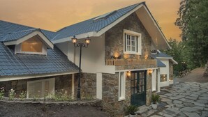 A two-story stone house with a steeply pitched roof, a front porch with a railing, and a small balcony with a wooden fence. - Echor Backwoods Villa Narkanda (Kumharsain)