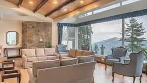 A spacious living room with a stone wall, large windows, and a view of mountains. - Echor Backwoods Villa Narkanda (Kumharsain)