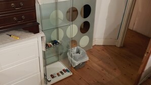 Ett rum med trägolv, en vit byrå med lådor, ett glasskåp med cirkelformade utskärningar och en papperskorg.